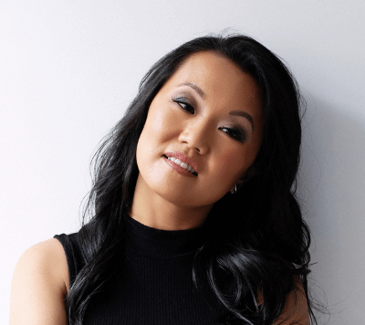 Against a white background, an Asian woman with long dark hair looks toward the camera with a slight smile, her head tilted to the right.