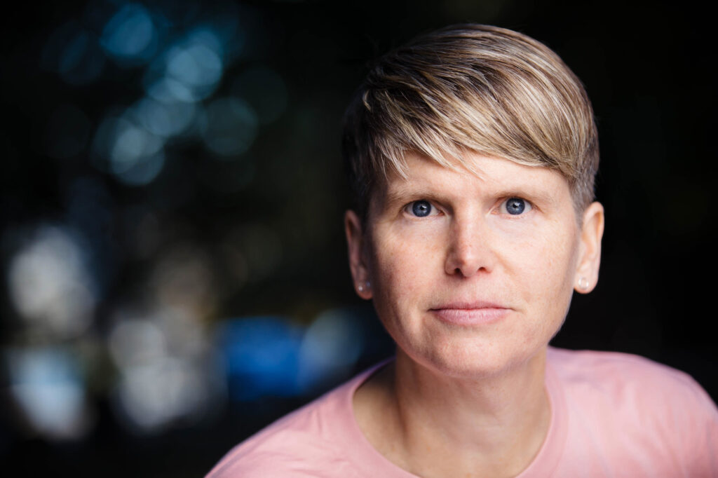 Against a dark backdrop, a white woman with short blonde hair stares intently into the camera. She wears a light pink shirt.