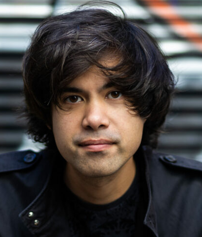 Against a graffitied background, a person with dark, somewhat shaggy hair, looks into the camera and has their hands crossed on a railing in front of them