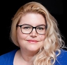 With a black background a woman with long, blonde, curly hair and glasses, wearing a blue cardigan, looks into the camera with a slight smile.