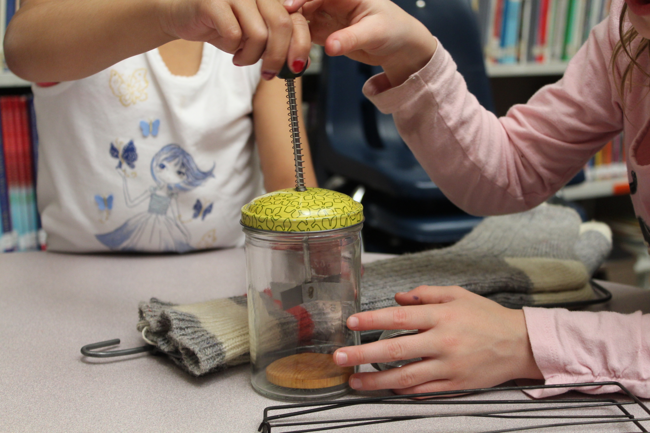 Two children holding a glass jar with a green lid and a wire wrapped straw. Only the arms and torsos of the children are visible.