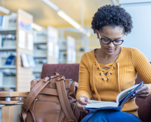 A woman sits looking at a book. A backpack is to the side and bookcases can be seen in the background.